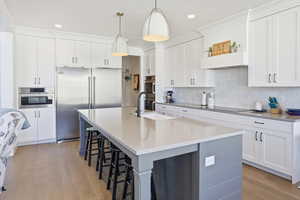 Kitchen featuring backsplash, appliances with stainless steel finishes, a kitchen island with sink, light wood finished floors, and white cabinetry