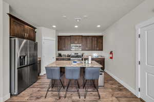 Kitchen featuring stainless steel appliances, light stone countertops, dark brown cabinetry, a kitchen breakfast bar, and dark wood finished floors