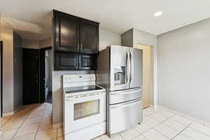 Kitchen with white electric range oven, light tile patterned flooring, stainless steel fridge, dark cabinets, and recessed lighting