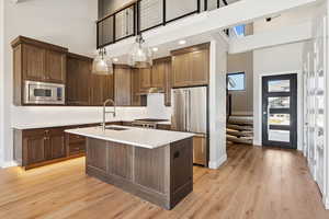 Kitchen featuring a towering ceiling, appliances with stainless steel finishes, hanging light fixtures, a center island with sink, and light wood-style floors