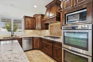 Kitchen with appliances with stainless steel finishes, tasteful backsplash, light stone counters, custom range hood, and dark brown cabinets