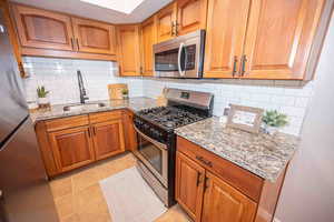 Kitchen featuring light stone countertops, stainless steel appliances, backsplash, brown cabinetry, and light tile patterned flooring