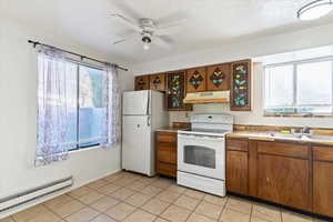 Kitchen featuring white appliances, baseboard heating, light tile patterned floors, under cabinet range hood, and brown cabinetry