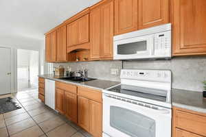 Kitchen featuring white appliances, tasteful backsplash, light tile patterned floors, brown cabinetry, and open shelves