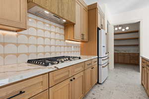 Kitchen with tasteful backsplash, custom exhaust hood, high end white refrigerator, light stone countertops, and open shelves