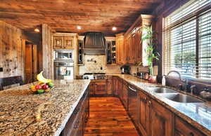 Kitchen featuring light stone counters, stainless steel appliances, custom exhaust hood, brown cabinetry, and recessed lighting
