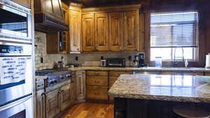 Kitchen featuring light stone counters, backsplash, stainless steel appliances, brown cabinetry, and custom exhaust hood