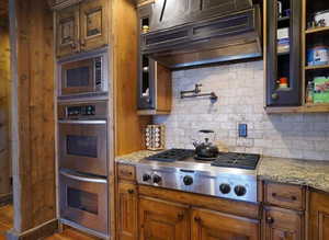 Kitchen with stainless steel appliances, custom exhaust hood, light stone countertops, decorative backsplash, and brown cabinetry