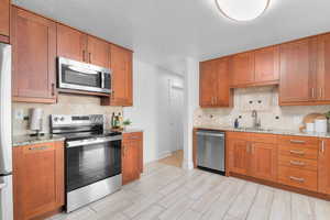 Kitchen featuring appliances with stainless steel finishes, backsplash, brown cabinetry, light stone counters, and a textured ceiling