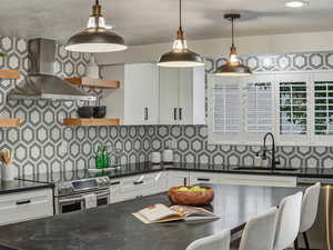 Kitchen with appliances with stainless steel finishes, white cabinetry, wall chimney range hood, pendant lighting, and a textured ceiling
