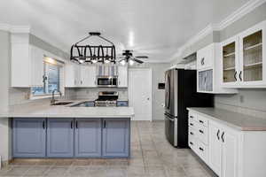 Kitchen featuring white cabinetry, blue cabinetry, light countertops, ornamental molding, appliances with stainless steel finishes, and a peninsula