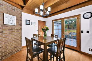 Dining room with brick wall, light wood-style flooring, a chandelier, and a wood ceiling with exposed beams