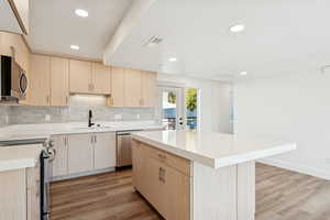 Kitchen featuring light brown cabinets, stainless steel appliances, light wood-style floors, backsplash, and a center island