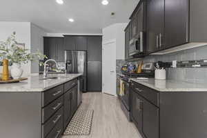 Kitchen featuring appliances with stainless steel finishes, light stone counters, light wood finished floors, backsplash, and a center island with sink