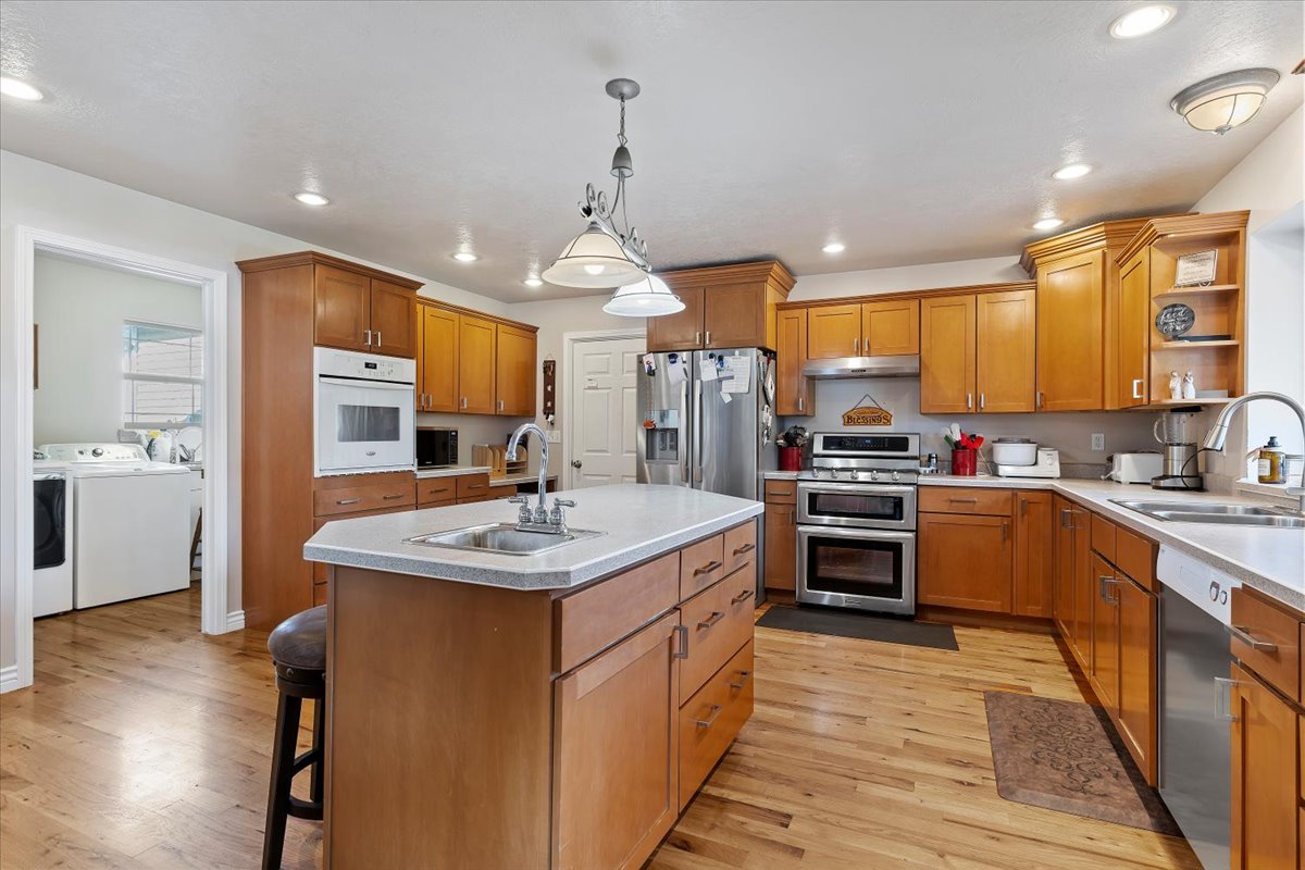 Kitchen with open shelves, brown cabinets, stainless steel appliances, hanging light fixtures, and an island with sink
