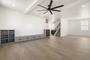 Unfurnished living room featuring beamed ceiling, light wood-style flooring, stairway, ceiling fan, and wooden walls