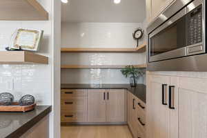Kitchen featuring open shelves, stainless steel microwave, light wood-style floors, light brown cabinets, and dark stone counters