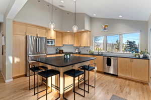 Kitchen featuring light brown cabinetry, a kitchen bar, appliances with stainless steel finishes, decorative light fixtures, and high vaulted ceiling