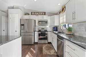 Kitchen with dark stone countertops, appliances with stainless steel finishes, vaulted ceiling, dark wood-style flooring, and decorative backsplash