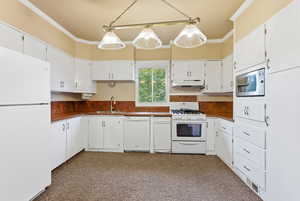 Kitchen featuring white appliances, dark countertops, crown molding, white cabinets, and under cabinet range hood