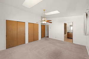 Unfurnished bedroom with a skylight, two closets, light colored carpet, ensuite bath, and a ceiling fan