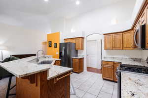 Kitchen featuring appliances with stainless steel finishes, brown cabinetry, a kitchen island with sink, light tile patterned floors, and light stone countertops