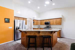 Kitchen with appliances with stainless steel finishes, a breakfast bar area, a kitchen island with sink, light carpet, and light stone counters