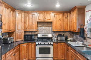 Kitchen featuring stainless steel range with gas stovetop, dark stone countertops, decorative backsplash, brown cabinets, and a textured ceiling