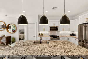 Kitchen featuring backsplash, light stone counters, a center island with sink, white cabinetry, and high end appliances