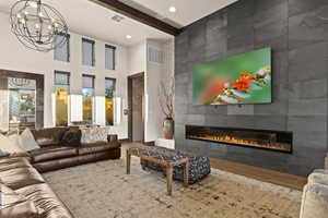 Living room with beam ceiling, a tiled fireplace, wood finished floors, a towering ceiling, and a chandelier