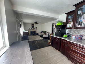 Kitchen featuring dark brown cabinetry, a fireplace with raised hearth, a textured ceiling, a ceiling fan, and light stone countertops
