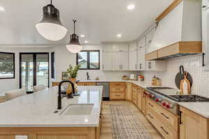 Kitchen featuring light brown cabinets, light stone countertops, tasteful backsplash, premium range hood, and appliances with stainless steel finishes