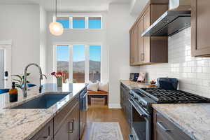 Kitchen featuring stainless steel appliances, wall chimney exhaust hood, light wood-type flooring, light stone counters, and backsplash