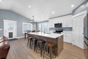 Kitchen with white cabinets, an island with sink, hanging light fixtures, appliances with stainless steel finishes, and vaulted ceiling