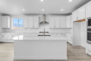 Kitchen featuring wall chimney exhaust hood, white cabinetry, recessed lighting, light wood-type flooring, and a center island with sink