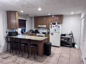Kitchen with a textured ceiling, white appliances, a kitchen breakfast bar, a peninsula, and light stone counters