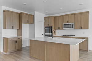 Kitchen featuring light stone counters, light wood-type flooring, appliances with stainless steel finishes, a center island with sink, and a textured ceiling