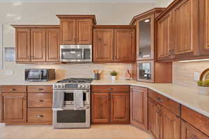 Kitchen featuring appliances with stainless steel finishes, brown cabinets, backsplash, light tile patterned floors, and glass insert cabinets