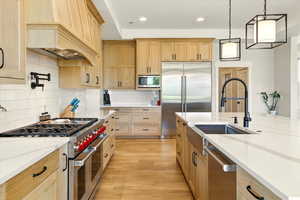 Kitchen featuring built in appliances, light brown cabinetry, tasteful backsplash, light stone countertops, and hanging light fixtures