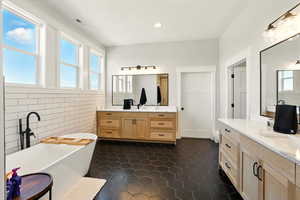 Full bath featuring a soaking tub, tile walls, two vanities, dark tile patterned flooring, and recessed lighting