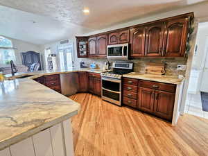 Kitchen featuring appliances with stainless steel finishes, decorative backsplash, light stone counters, light wood finished floors, and a textured ceiling