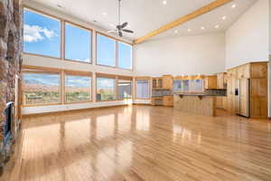 Unfurnished living room featuring light wood-style flooring, a ceiling fan, a high ceiling, a stone fireplace, and beamed ceiling