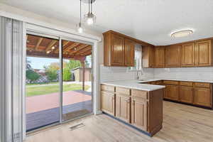 Kitchen featuring light wood-type flooring, tasteful backsplash, pendant lighting, brown cabinetry, and a textured ceiling
