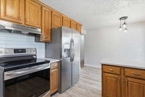 Kitchen featuring appliances with stainless steel finishes, brown cabinets, under cabinet range hood, decorative backsplash, and decorative light fixtures