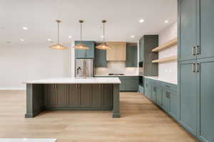 Kitchen with green cabinetry, an island with sink, backsplash, open shelves, and decorative light fixtures