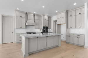 Kitchen with gray cabinetry, light stone counters, light wood-style flooring, a kitchen island with sink, and recessed lighting