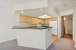 Kitchen featuring dark countertops, light colored carpet, light brown cabinetry, white appliances, and a textured ceiling