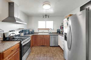 Kitchen with appliances with stainless steel finishes, wall chimney range hood, a textured ceiling, brown cabinetry, and light tile patterned floors
