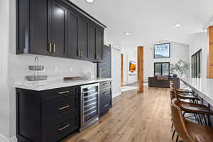 Indoor bar featuring light wood-style flooring, wine cooler, light stone countertops, recessed lighting, and dark cabinets