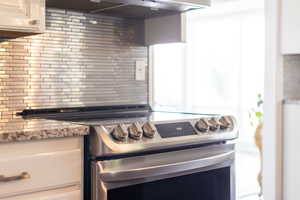 Kitchen with electric stove, decorative backsplash, range hood, and white cabinets